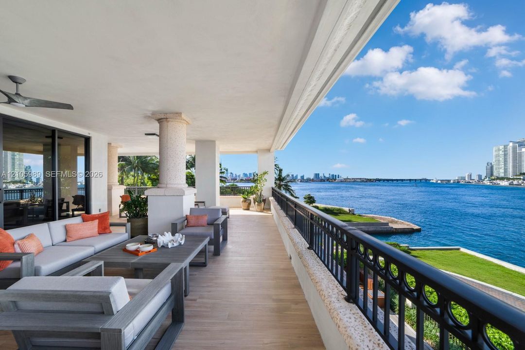Expansive covered balcony featuring wood-finish flooring, architectural columns, a black metal railing, and a ceiling-mounted fan, overlooking the water