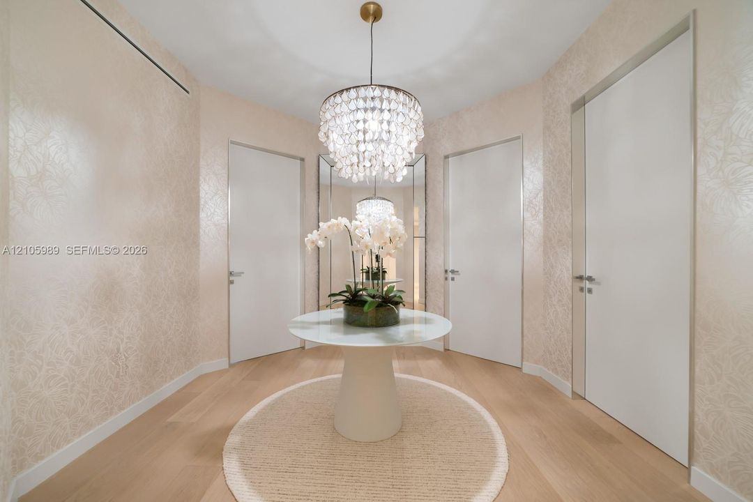Foyer featuring light-toned wood-finish flooring, textured wall coverings, a tiered crystal chandelier, full-height flush doors with metallic casing, and floor-to-ceiling mirror panels