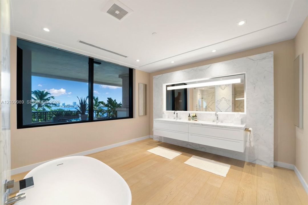 Floating dual vanity with integrated countertop, floor-to-ceiling stone wall treatment, back-lit mirror, freestanding soaking tub, and large-format black-framed windows