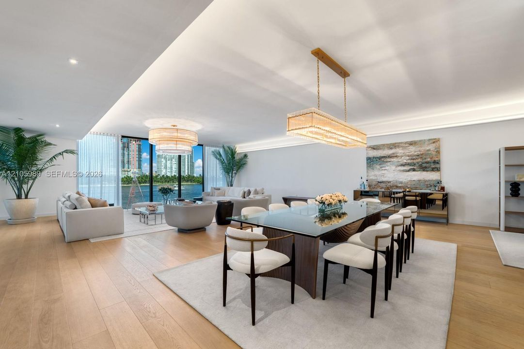 Open-concept living space featuring wood-finish flooring, recessed perimeter ceiling lighting, floor-to-ceiling windows, and dual statement chandeliers