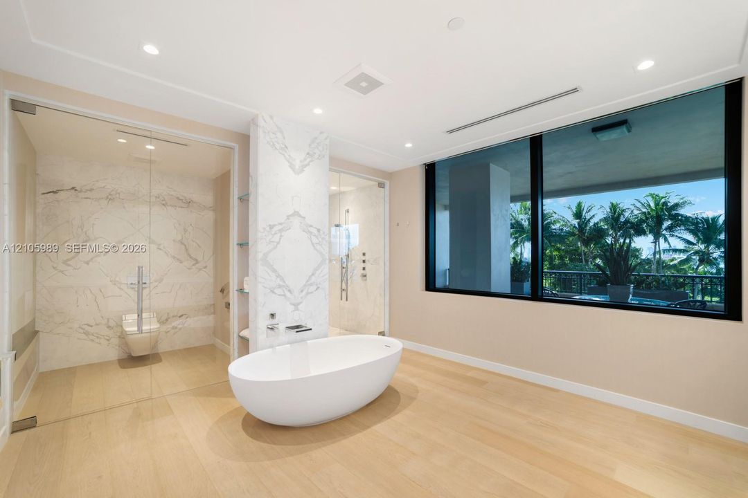Freestanding soaking tub, stone-clad feature wall, glass-enclosed shower, wood-finish flooring, and expansive window with exterior greenery