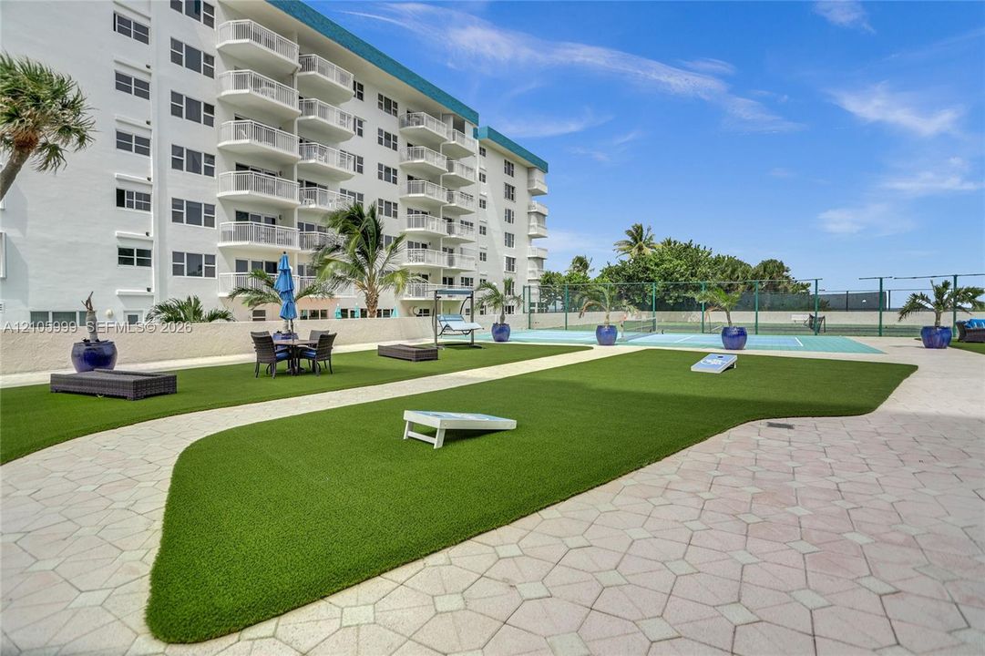 Outdoor recreation area featuring artificial turf, stone-paver hardscaping, a fenced tennis court, and high-rise building with private balconies
