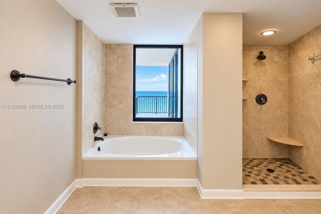 Deep soaking tub, walk-in shower with built-in bench seating, neutral stone-finish tile surround, dark bronze hardware fixtures, and exterior ocean backdrop