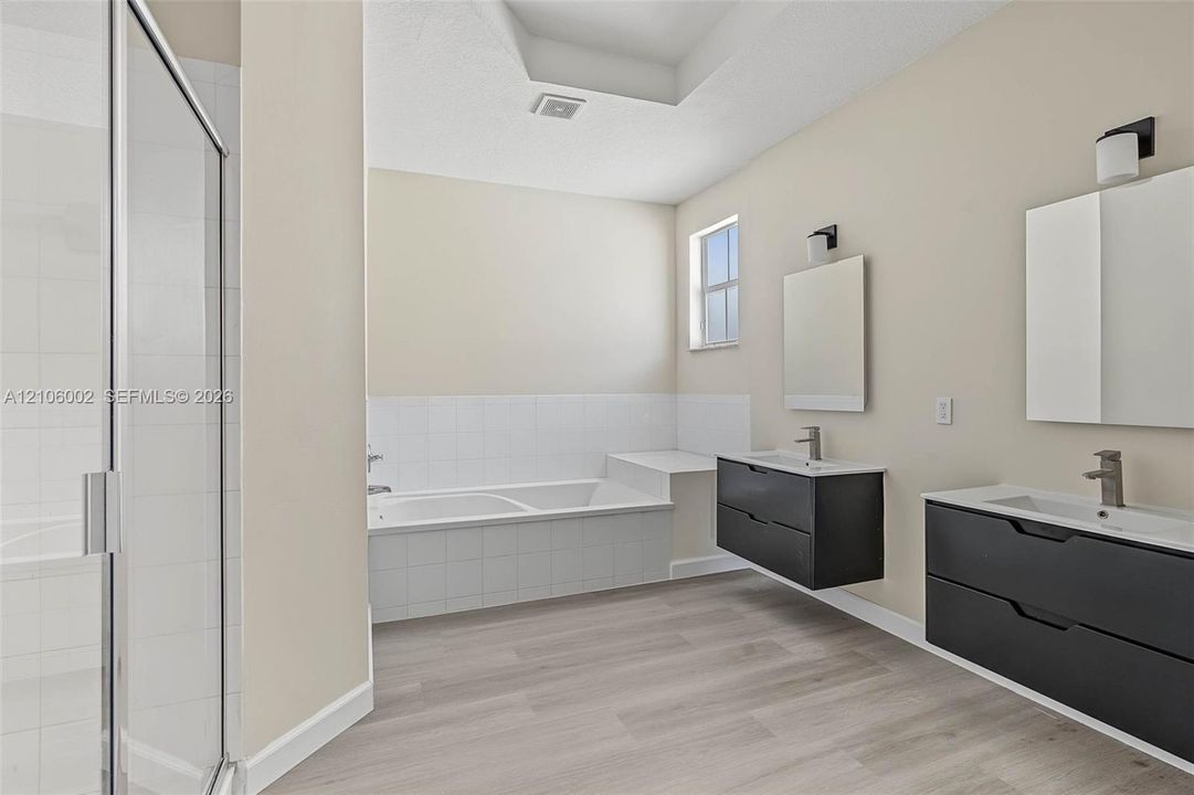 Bathroom featuring dual floating vanities with dark cabinetry, integrated white countertops, rectangular mirrors, and wood-finish flooring, complemented by a tiled soaking tub and a glass-enclosed shower