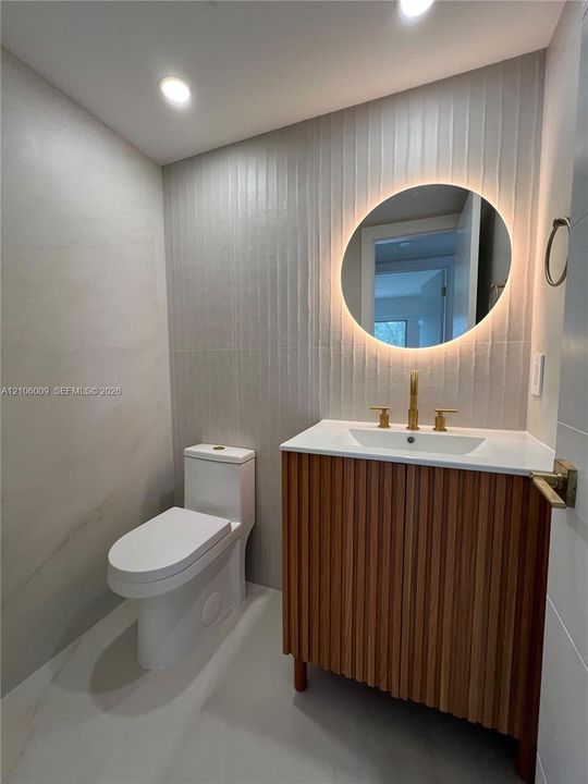 Modern vanity with wood-finish vertical slats, white countertop, integrated sink, and brass-finish faucet, complemented by a backlit circular mirror, textured vertical wall tile, and recessed lighting