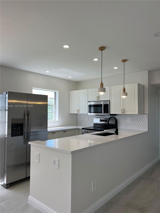 Kitchen featuring white shaker-style cabinetry, white solid-surface countertops, stainless steel appliances, a hexagonal tile backsplash, and two minimalist pendant lights