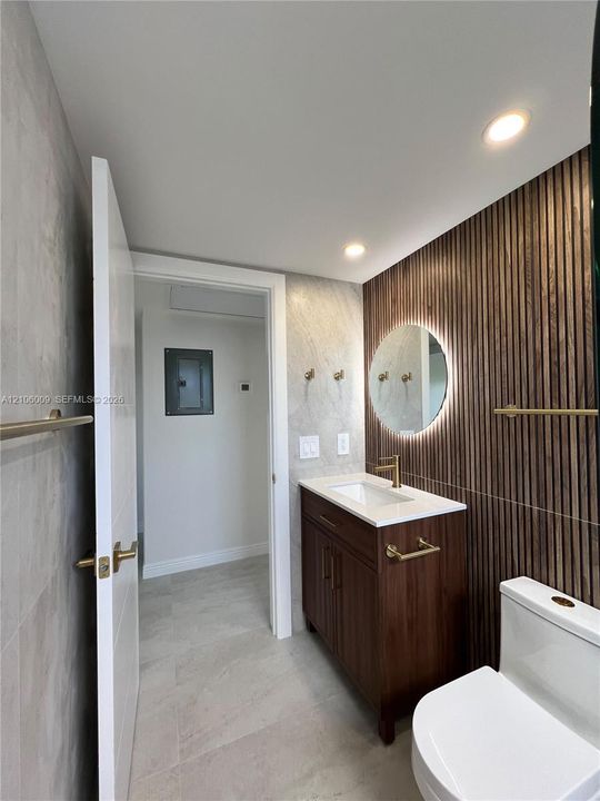 Wood-slat accent wall featuring a backlit circular mirror, vanity with a white countertop, and gold-finish hardware throughout