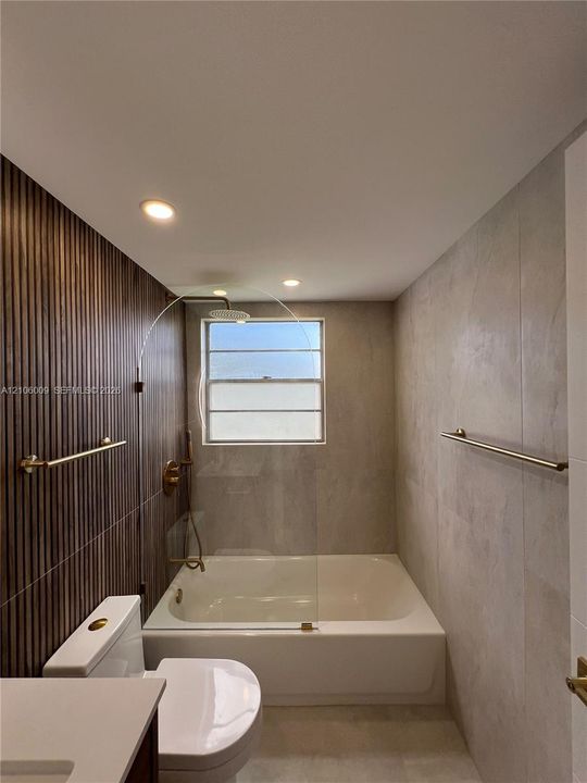 Bathtub featuring a frameless glass panel, vertical wood-slat feature wall, large-format tile surround, recessed lighting, and brass-finish plumbing fixtures