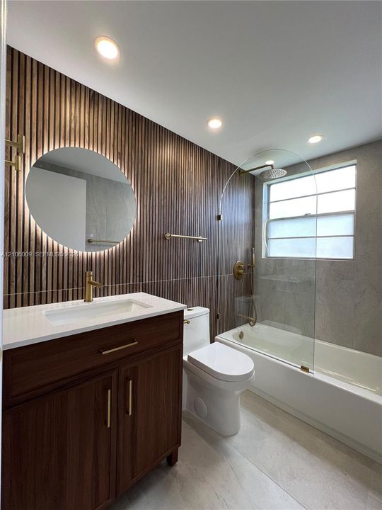 Bathroom featuring a wood-slat accent wall, circular LED-backlit mirror, brass-finish fixtures, wooden vanity with a solid-surface countertop, and a glass-enclosed tub with stone-finish tile surround