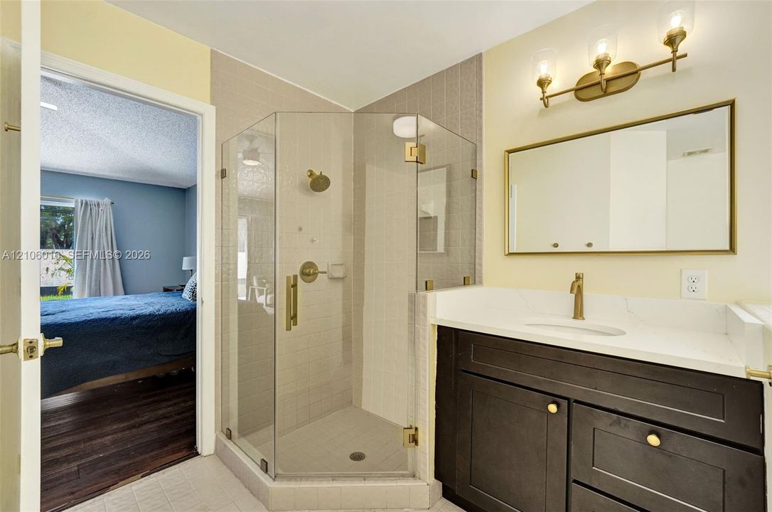Bathroom featuring a corner glass-enclosed shower, dark wood-finish vanity with a light stone-style countertop, rectangular mirror with gold-tone frame, and matching gold-tone plumbing fixtures