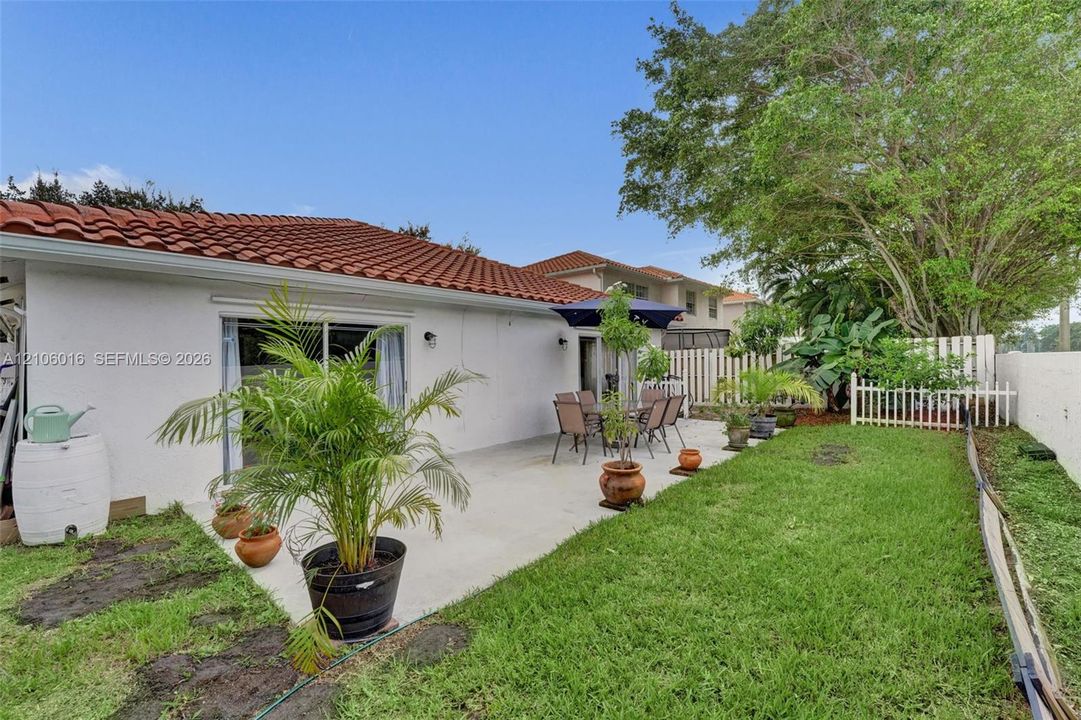 Rear exterior featuring a concrete patio, white stucco siding, a terra-cotta tile roof, and a fenced grass lawn with mature trees
