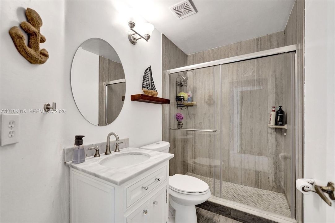 Bathroom featuring a white vanity with a stone countertop, an oval wall-mounted mirror, and a walk-in shower with sliding glass doors and large-format tile surround