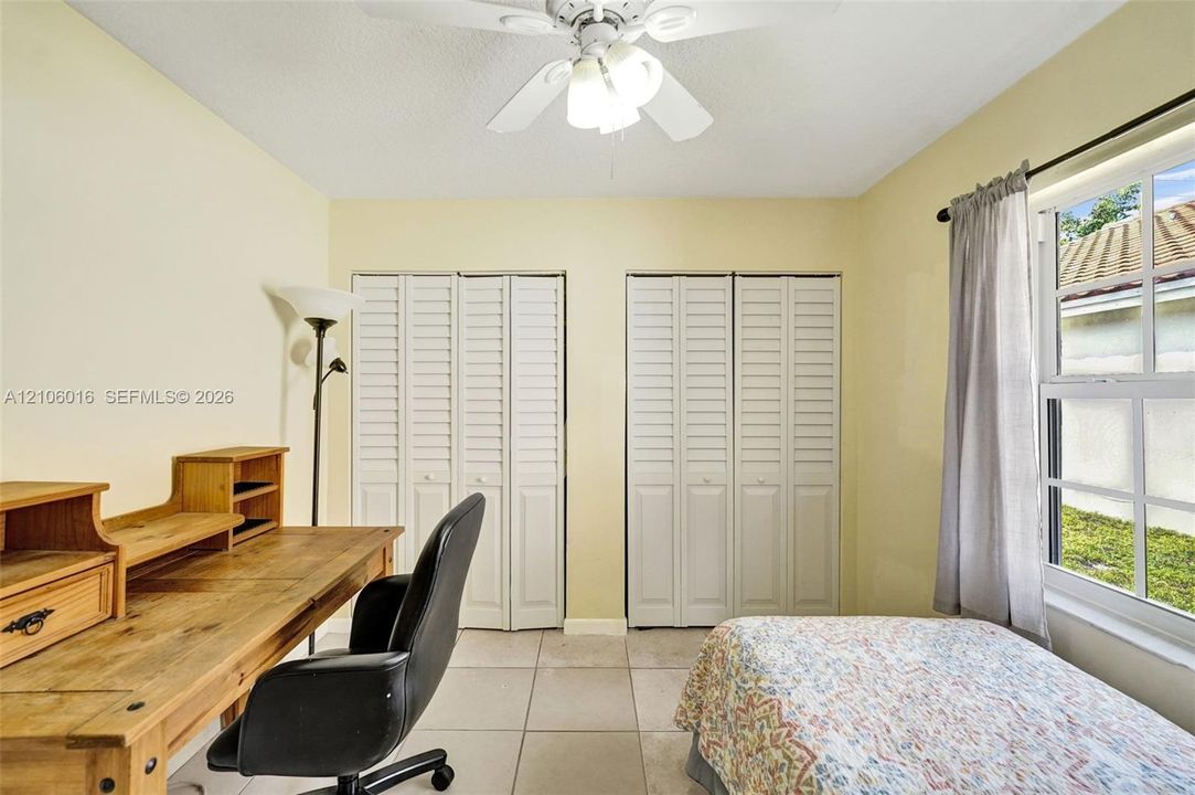 Interior room featuring light-toned ceramic tile flooring, two sets of white louvered-style bi-fold closet doors, a multi-blade ceiling fan with integrated lighting, and a single-hung window with a metal rod and drapery hardware