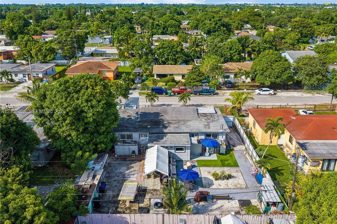 Aerial perspective of a detached residential property featuring a flat roofline, fenced perimeter, concrete-surfaced rear yard, synthetic turf area, and mature shade trees