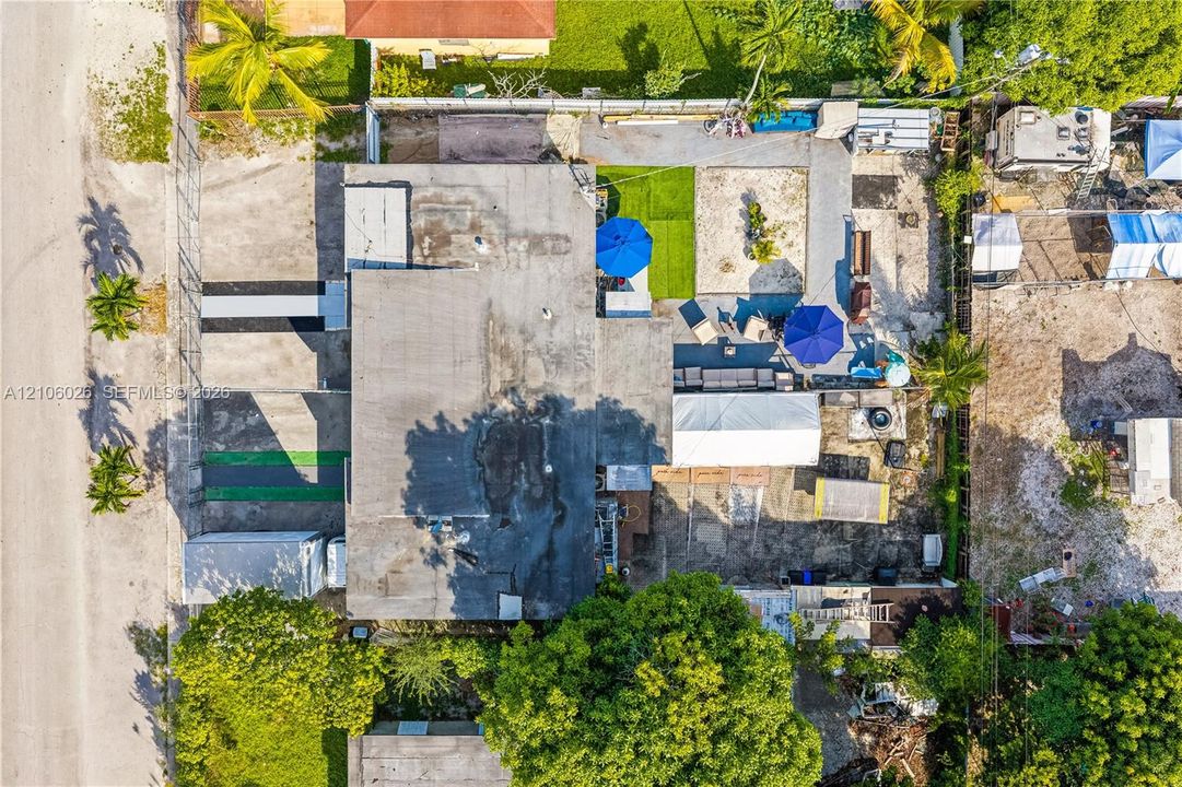Aerial perspective of a property featuring a concrete-surface roof, paved driveway, partitioned exterior lot sections, and mature perimeter trees