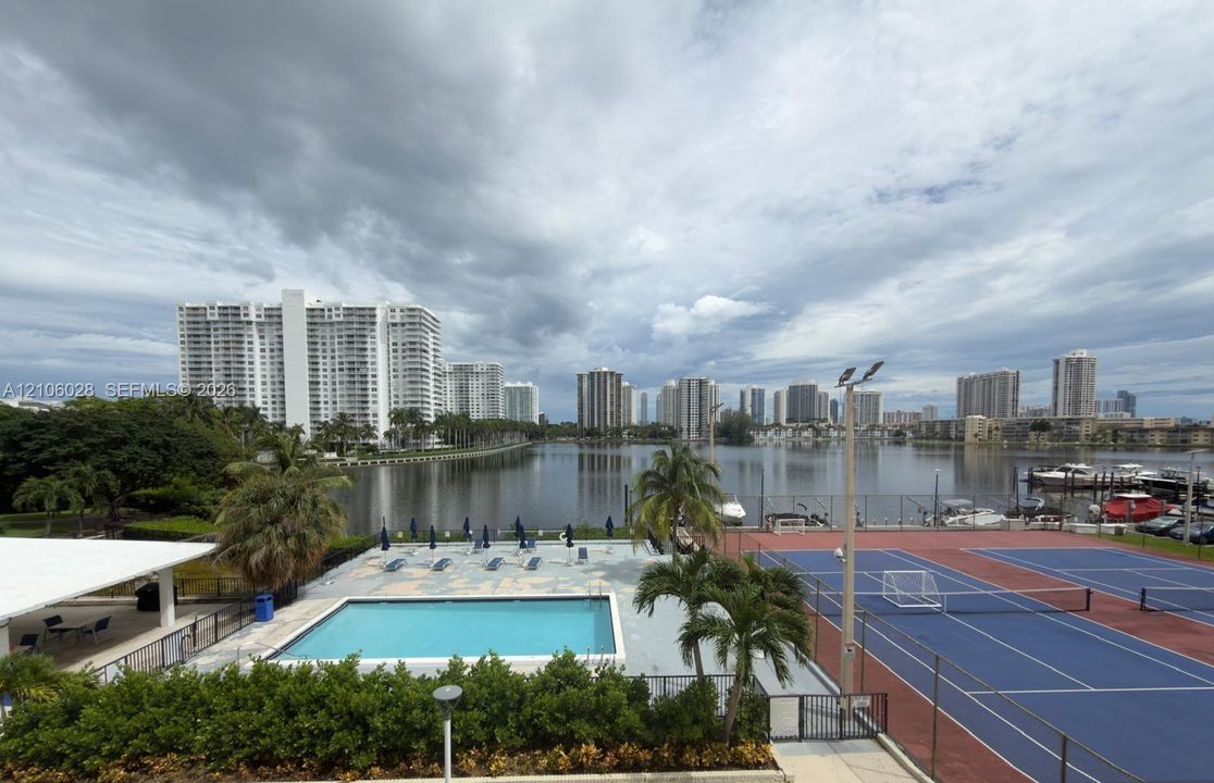 Community recreational area featuring a swimming pool, hard-court tennis facilities, and landscaped palm trees, positioned alongside a waterfront with boat docking