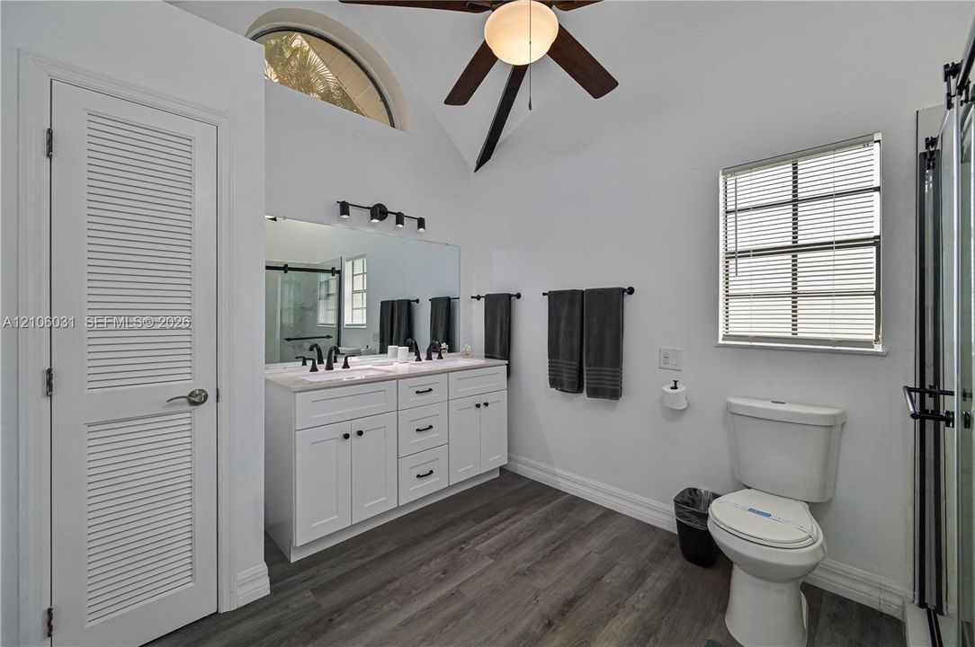 Double vanity with white cabinetry, neutral stone-finish countertop, dual under-mount sinks, and matte black hardware, featuring wood-finish flooring, vaulted ceilings, and a fixed arched window