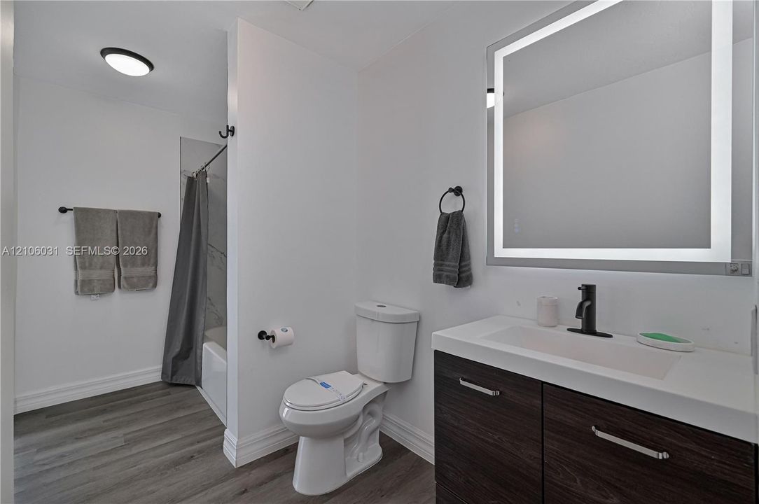 Bathroom featuring a dark wood-finish vanity with a rectangular integrated sink, matte black faucet, illuminated wall mirror, and grey wood-finish flooring