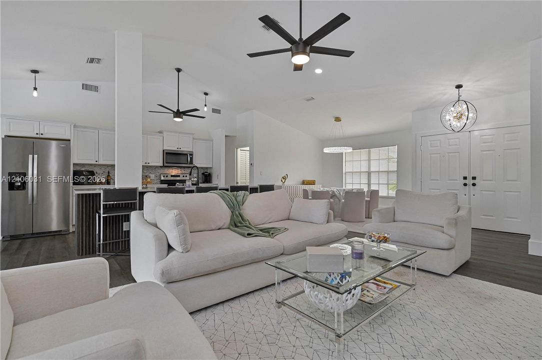 Open-concept interior featuring vaulted ceilings, wood-finish flooring, white shaker cabinetry, stainless steel kitchen appliances, and contemporary multi-arm chandeliers