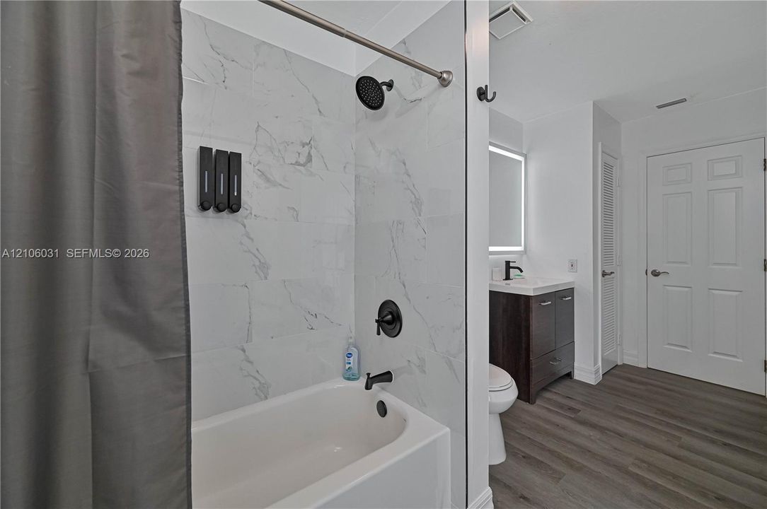Bathroom featuring a tub with large-format light-toned tile surround, matte black shower fixtures, wood-finish vanity with a rectangular sink, illuminated mirror, and wood-finish flooring