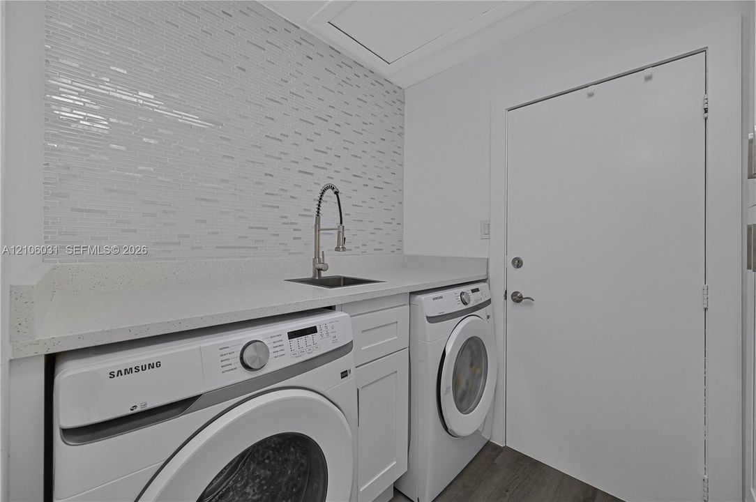 Laundry area featuring a stone countertop, an integrated sink with a gooseneck faucet, a textured tile backsplash, white shaker-style cabinetry, and wood-finish flooring