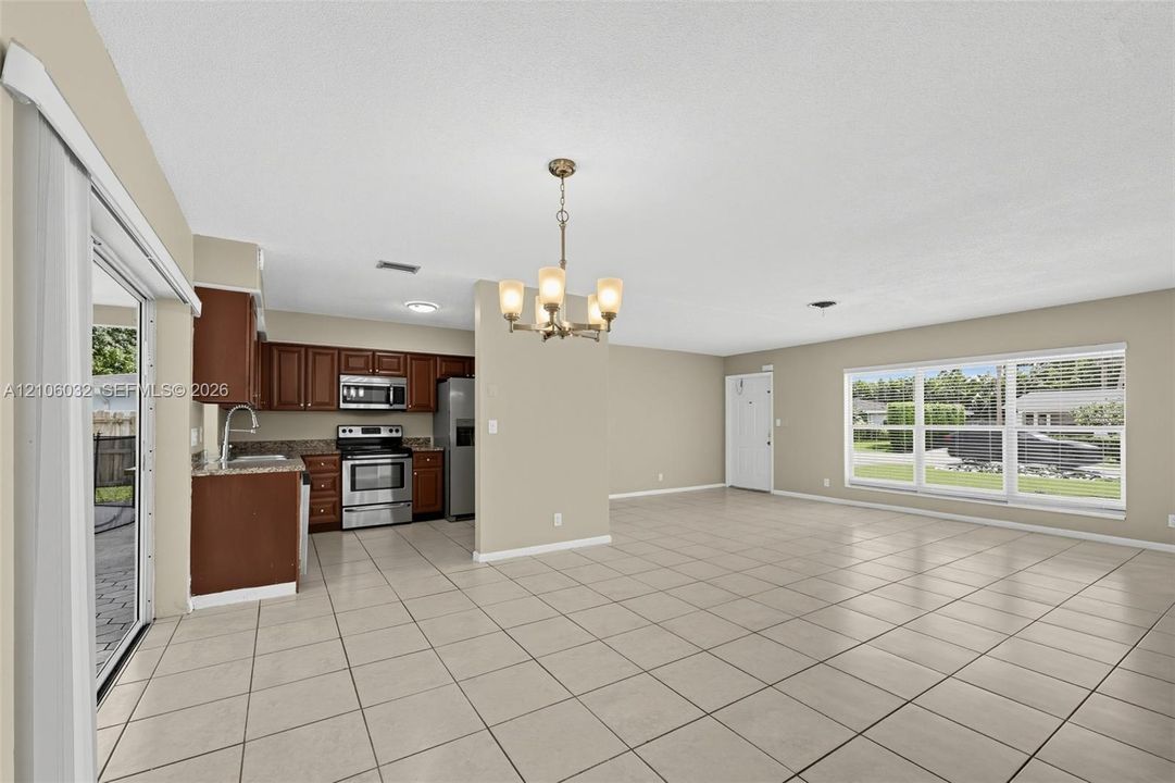 Open-concept interior featuring light-toned tile flooring, stainless steel appliances, dark wood cabinetry, a multi-light chandelier, and large windows