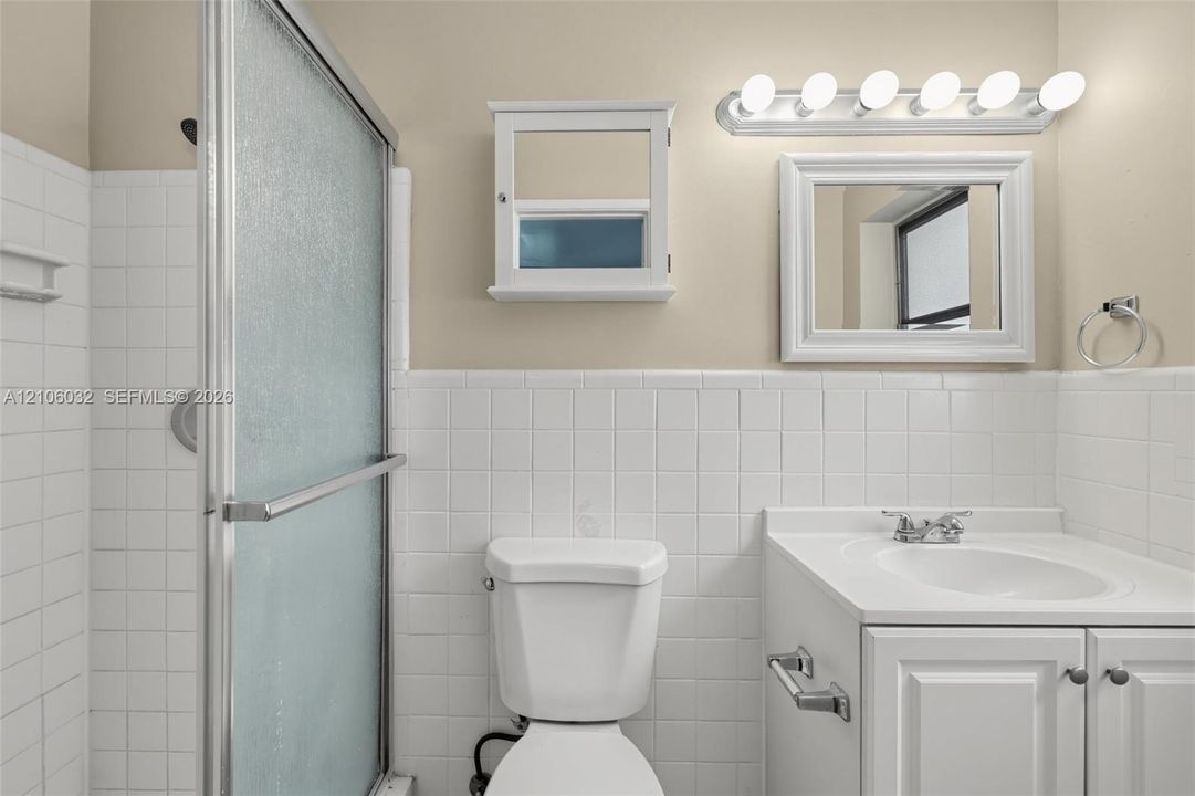 Bathroom featuring white square tile wainscoting, shower enclosure with frosted glass panels, white vanity cabinet with a solid-surface top, wall-mounted medicine cabinet, and six-bulb vanity lighting
