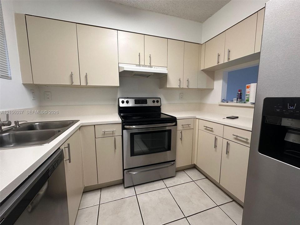 Kitchen featuring cream-toned cabinetry with metal hardware, solid-surface countertops, stainless steel appliances, a dual-basin sink, and tile flooring