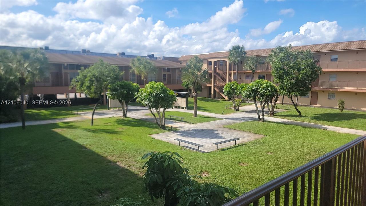 Exterior FRONT VIEW courtyard featuring manicured lawn, paved pathways, mature trees, and ground-level seating