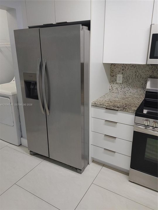 Stainless steel side-by-side refrigerator, white flat-panel cabinetry, stone-surface countertops, and large-format tile flooring