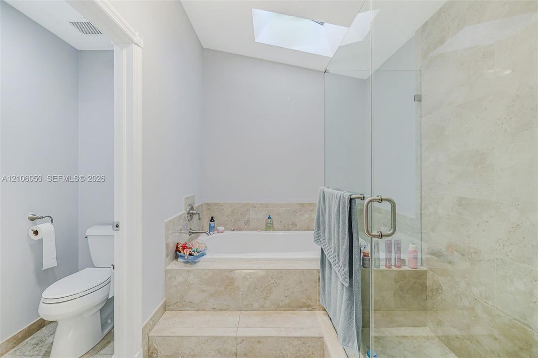 Skylit bathroom featuring a tiled platform soaking tub, separate glass-enclosed shower, stone-finish surround, and a porcelain toilet