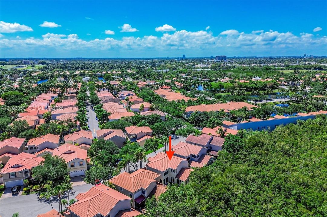 Aerial perspective of a multi-story residence featuring a tile roof, light-toned exterior finish, and a concrete driveway situated within a landscape of established trees and vegetation