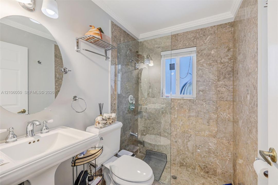 Pedestal sink with chrome faucet, oval wall-mounted mirror, and walk-in shower featuring glass enclosure and natural stone tile surround