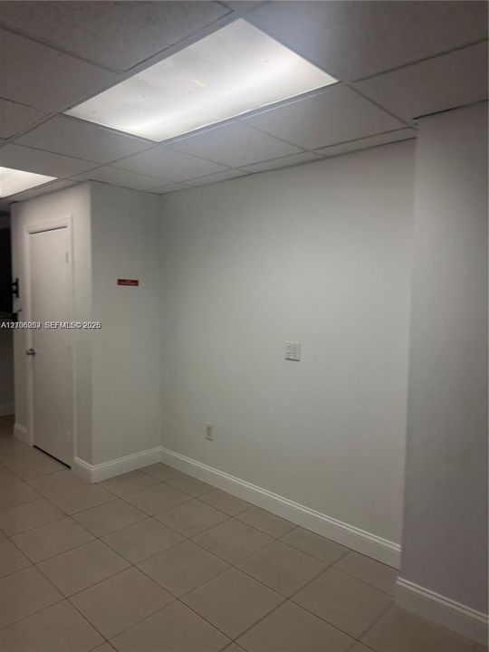 Neutral-toned interior space featuring grid-pattern drop ceiling, recessed lighting panels, ceramic tile flooring, and solid-core interior door