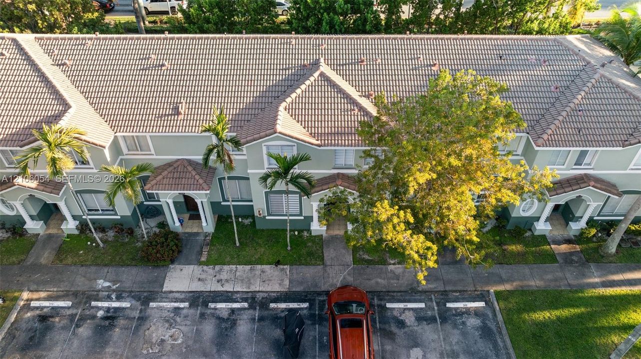 Multi-unit building exterior featuring a tile roof, light green stucco facade, decorative columns, arched entryways, and manicured palm trees
