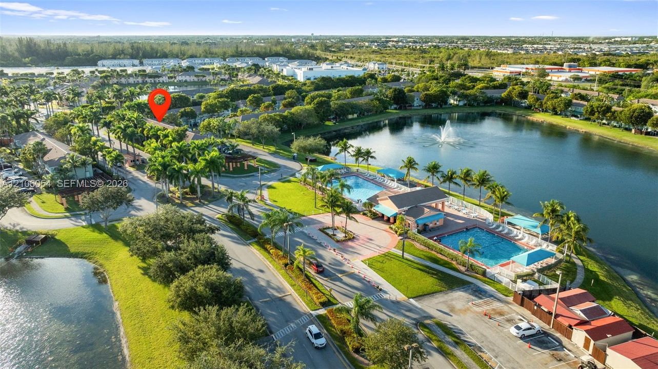 Aerial perspective of a community featuring a large retention pond, a central fountain, two swimming pools, and extensive palm tree landscaping