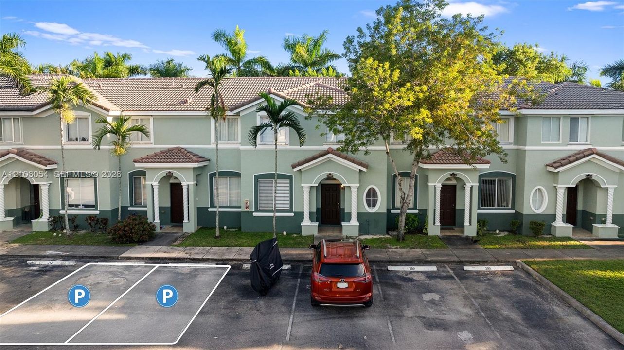 Two-story townhome exterior featuring a light green and dark green facade, tiled gable roofline, arched entryways with decorative columns, and assigned surface parking spaces