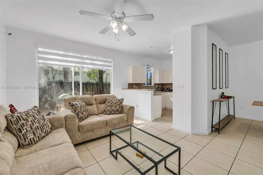 Living area featuring large sliding glass doors, neutral tile flooring, ceiling fan with light kit, and pass-through counter to kitchen