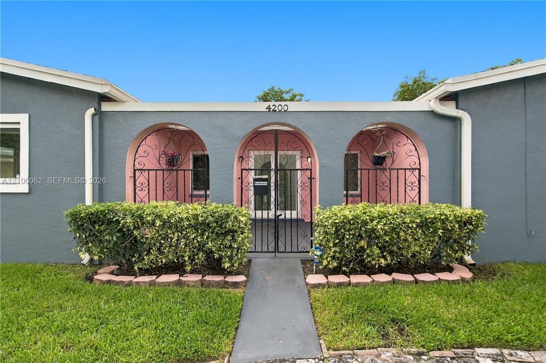Stucco exterior featuring a triple-arched entryway, decorative wrought-iron security gate, and low-profile shrubbery lining the concrete walkway