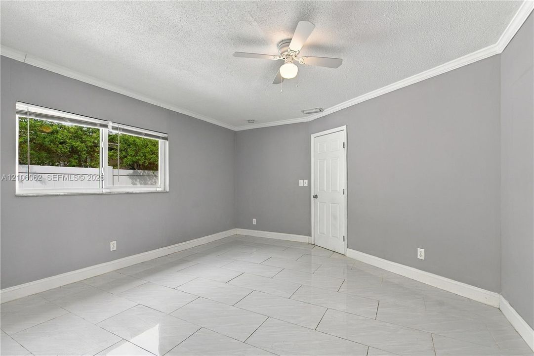 Neutral-toned interior featuring large-format tile flooring, a centered ceiling fan with integrated lighting, crown molding, and a single-panel interior door