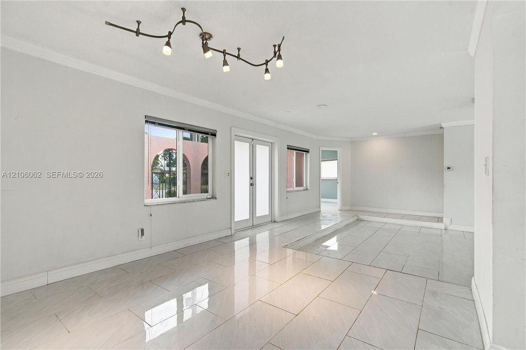 Neutral-toned interior featuring large-format tile flooring, multi-light track lighting, white crown molding, double glass-panel entry doors, and a sunken floor section