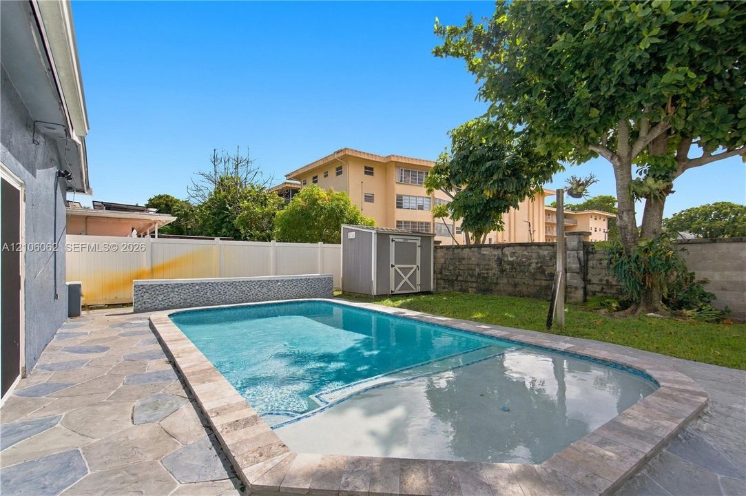 In-ground swimming pool with a shallow sun shelf, stone-tile decking, decorative tiled retaining wall, and a detached storage shed