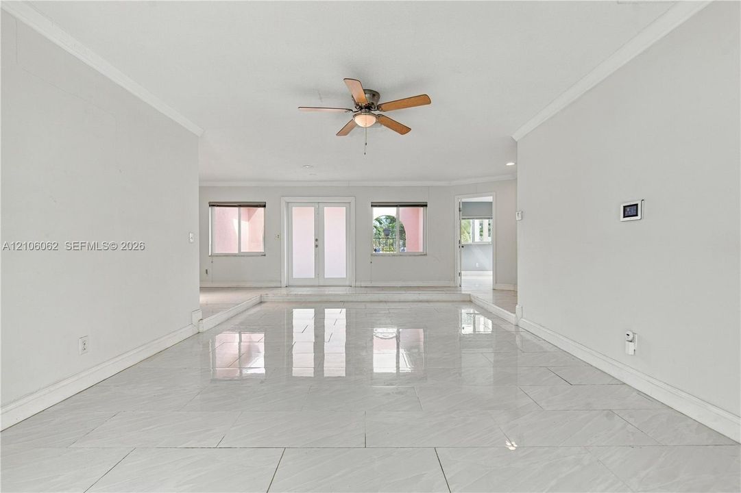 Open-concept interior featuring large-format tile flooring, crown molding, a central ceiling fan, glass-paned double doors, and a recessed step-down floor detail