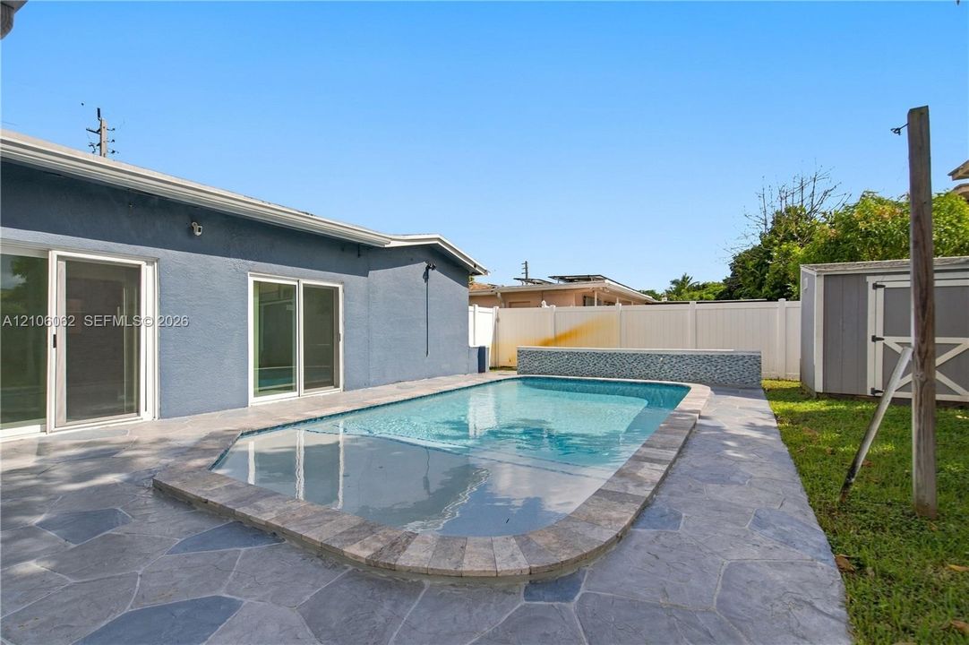 In-ground swimming pool with stone coping and mosaic tile detail, flagstone patio, and exterior utility shed enclosed by white vinyl fencing