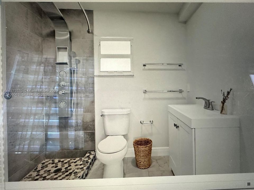 Walk-in shower featuring wall-mounted body sprayers, glass enclosure, and pebble-style stone flooring, complemented by a white vanity with integrated basin and large-format tile wall cladding