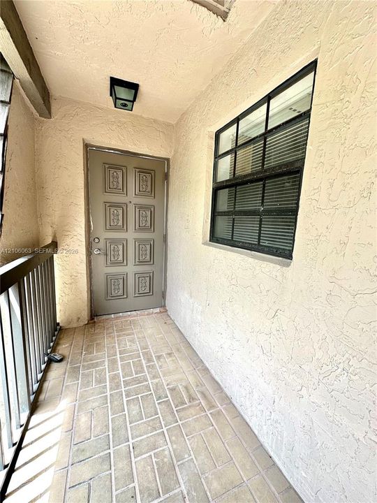 Covered exterior entry featuring textured stucco walls, paved brick-pattern flooring, a paneled metal door with decorative molding, and a multi-pane window with black framing