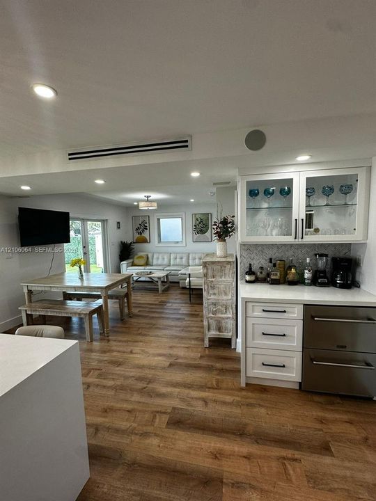 Custom built-in bar featuring white shaker-style cabinetry, glass-front upper displays, stone-pattern backsplash, and stainless-steel under-counter refrigeration, all complemented by wood-finish flooring and recessed lighting