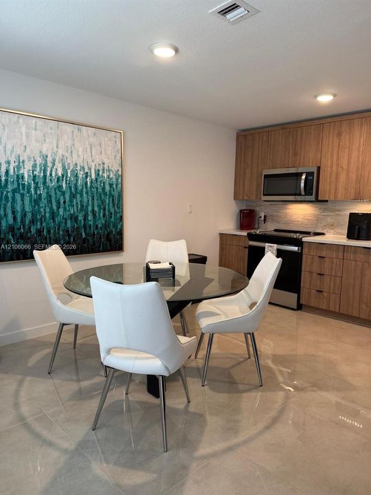 Kitchen featuring wood-finish cabinetry, stone-textured tile backsplash, light-colored countertops, stainless steel appliances, and large-format tile flooring