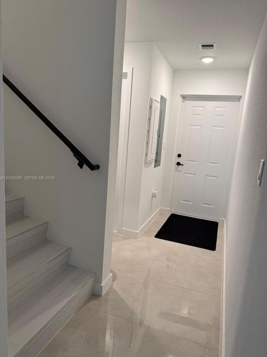 Entryway featuring large-format neutral tile flooring, a six-panel exterior door with matte black hardware, and a staircase with wood-finish treads and a matte black wall-mounted handrail