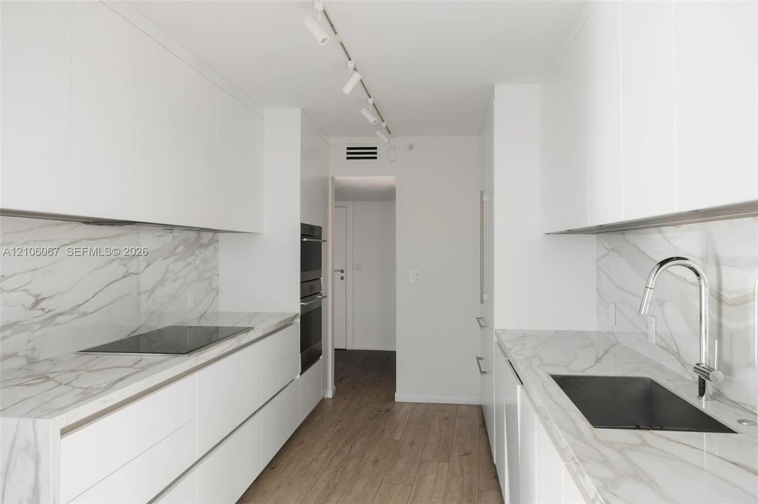 Galley kitchen featuring flat-panel white cabinetry, stone-surface countertops and backsplashes, built-in dual ovens, integrated cooktop, and wood-finish flooring